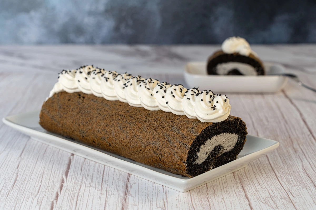 Chocolate keto swiss roll cake with black sesame and cream on a white plate against a blurred background